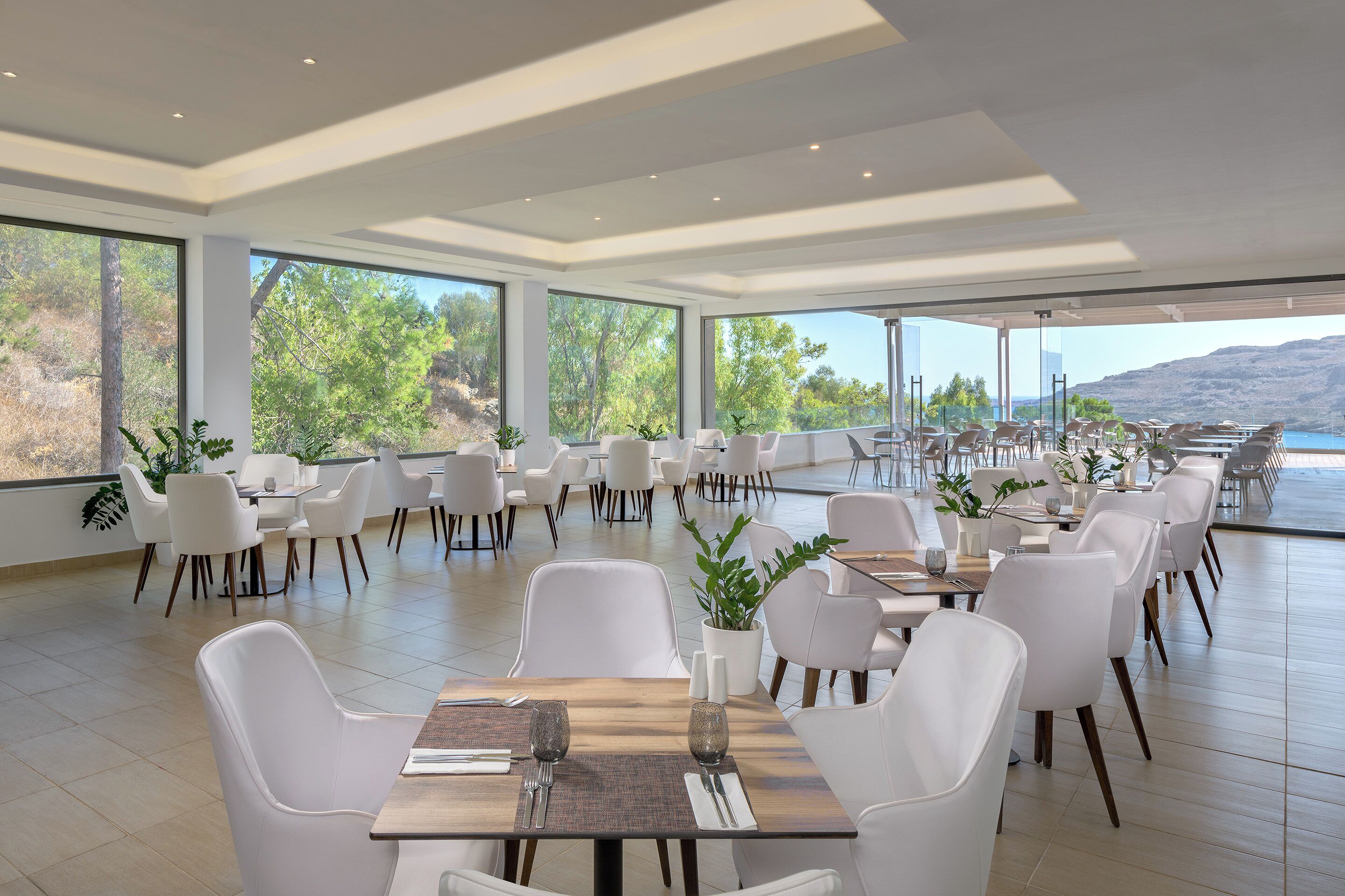 Une salle à manger spacieuse, avec des chaises et des tables blanches, de grandes fenêtres offrant une vue sur les arbres et un plan d’eau.