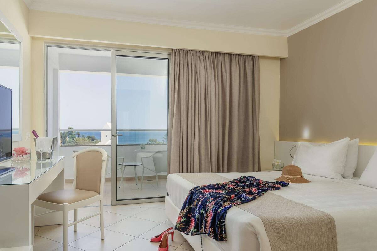 A hotel room with a bed, a desk, a chair, and a view of the sea through a sliding glass door.