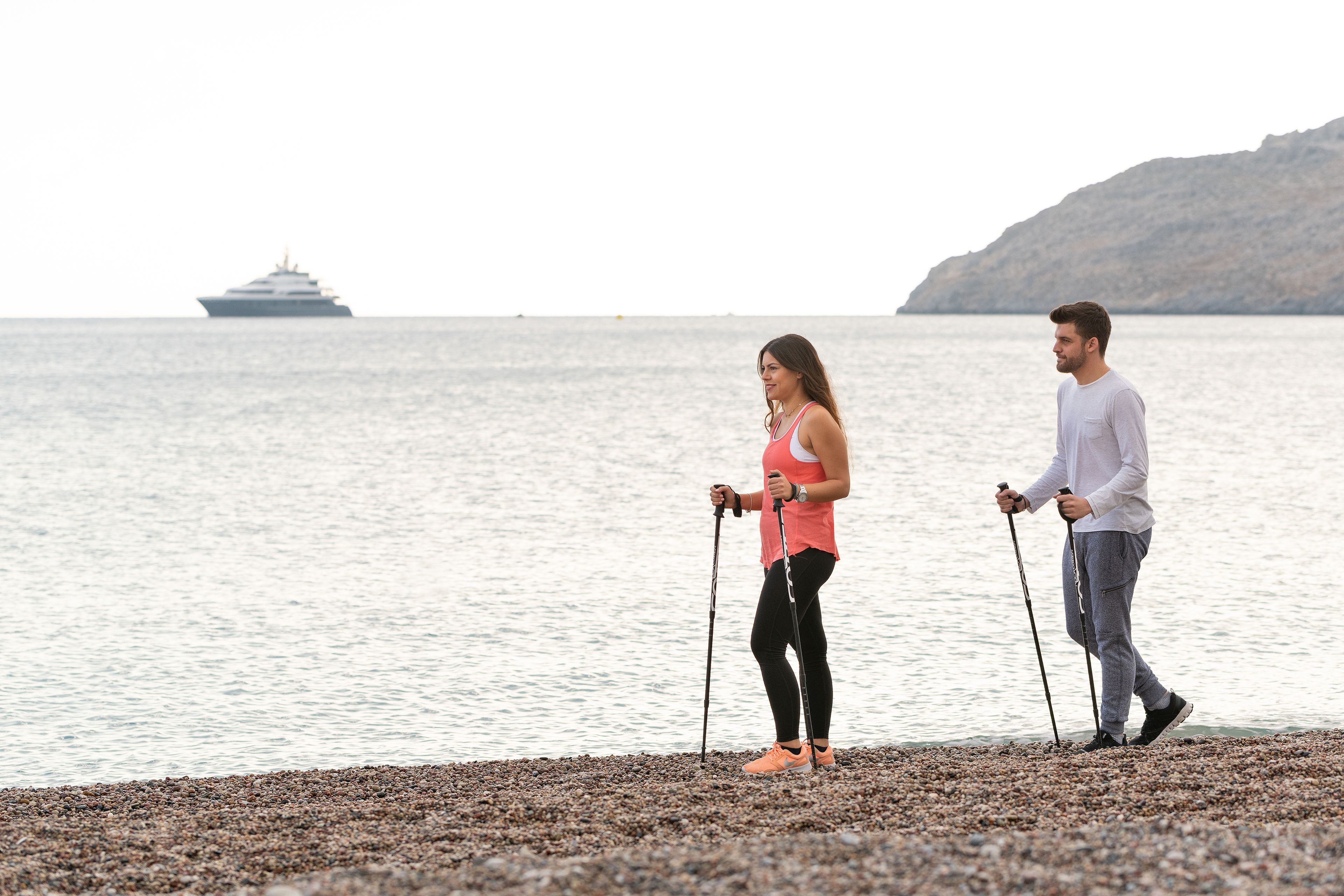 Two people walking on a pebbled beach with a ship in the background.