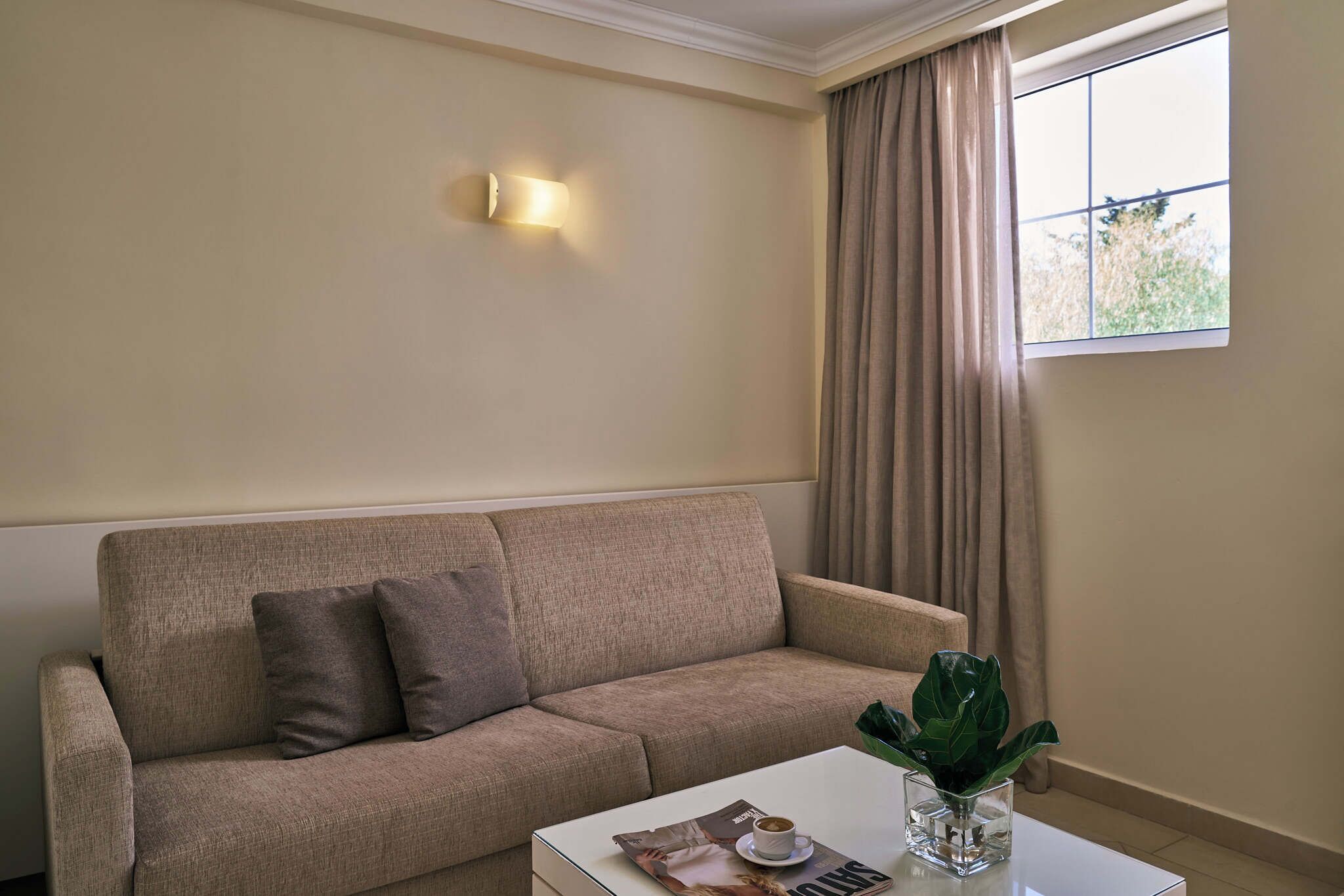 A living room with a beige sofa, a coffee table with a magazine and a plant, a wall-mounted light, and a window with curtains.