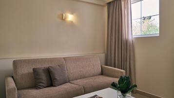 A living room with a beige sofa, a coffee table with a magazine and a plant, a wall-mounted light, and a window with curtains.