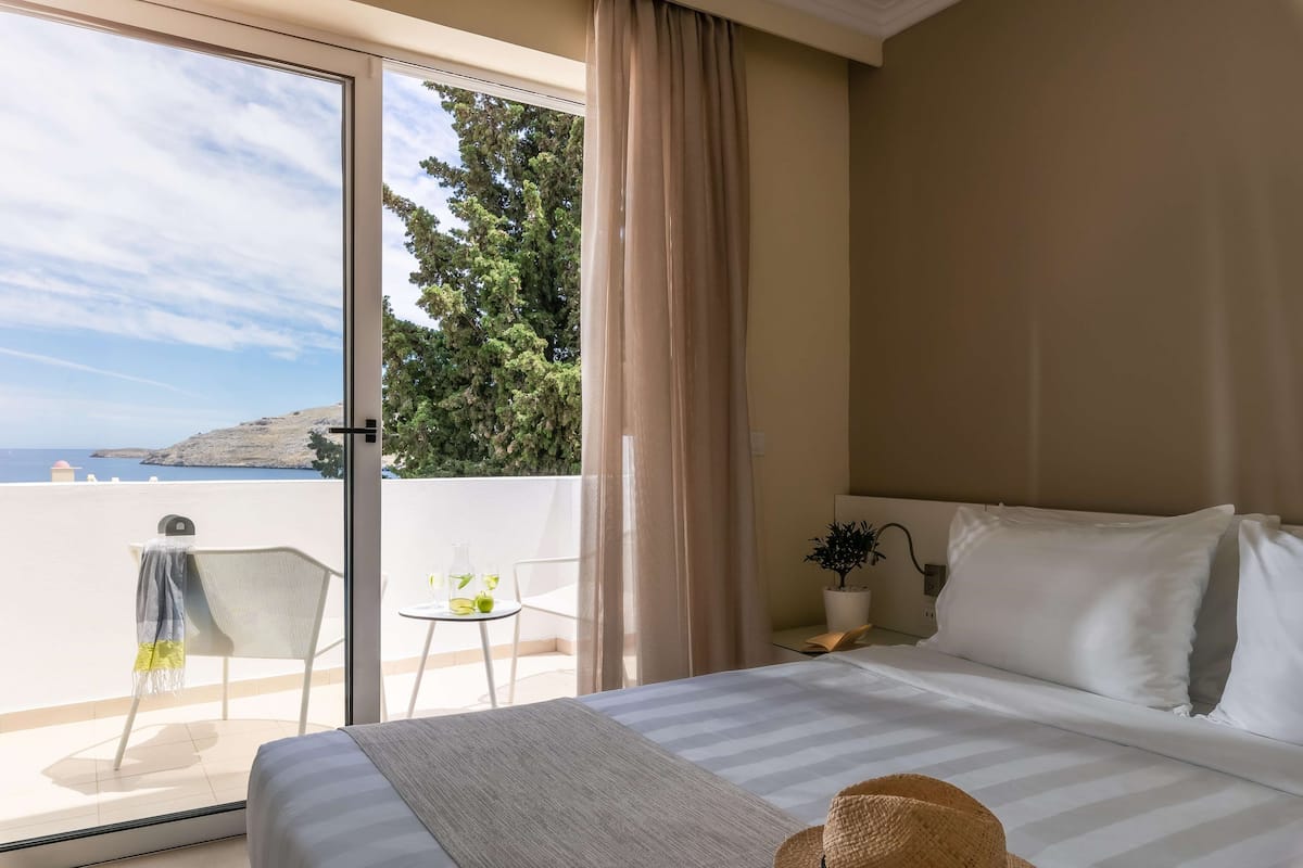 A hotel room with a bed, a small table with a chair, a potted plant, and a view of the sea and trees through a sliding glass door.