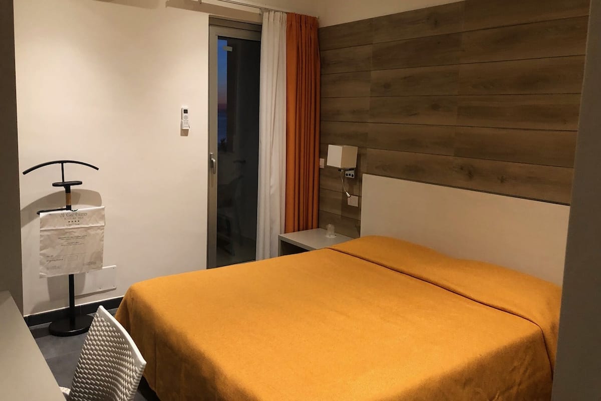 A hotel room with a bed, a desk, a chair, a nightstand, a lamp, a window with curtains, and an air conditioning unit.