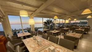Un restaurante con mesas dispuestas para comer, amplias ventanas con vista al océano e iluminación colgante.