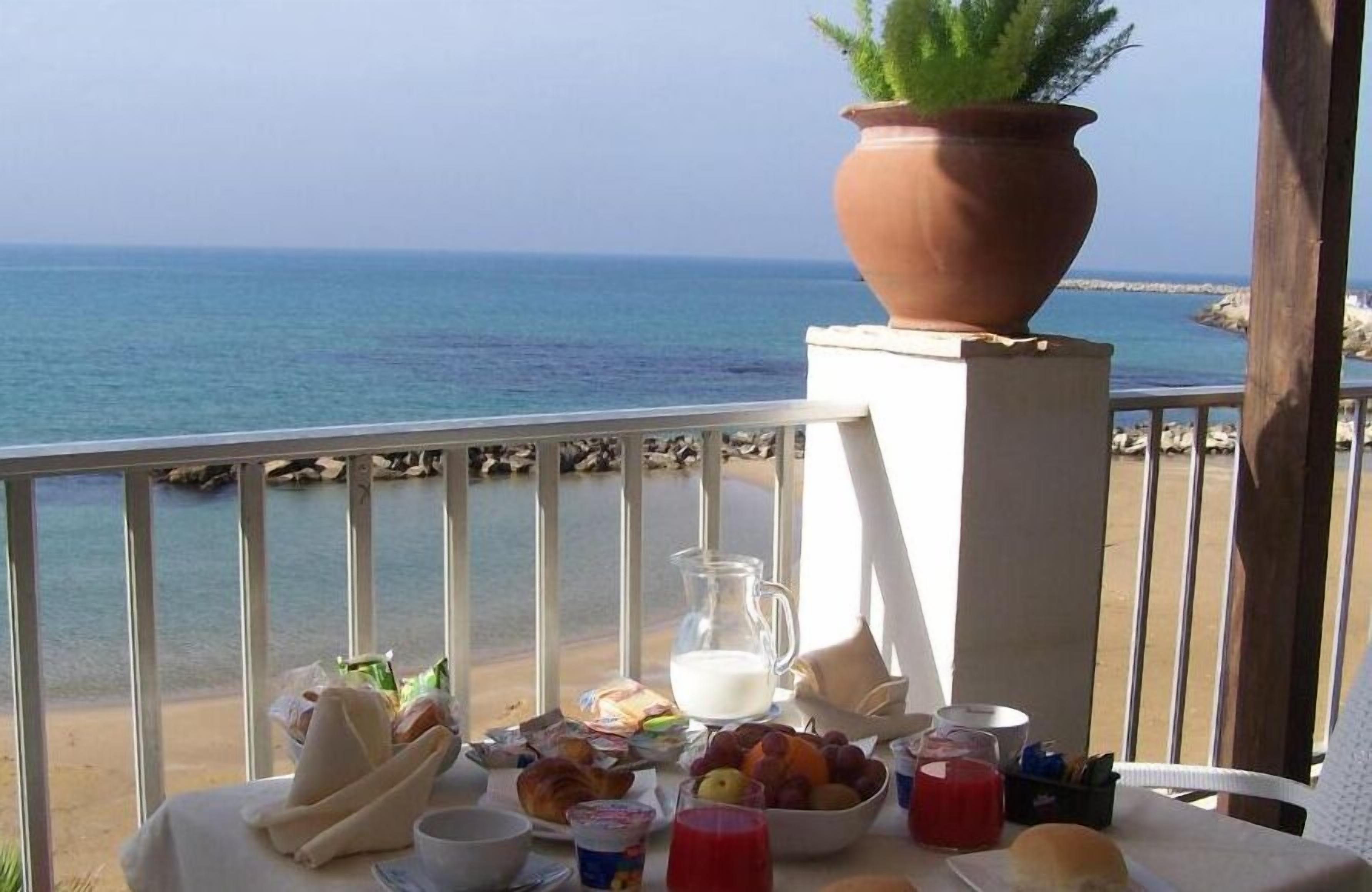 A table with breakfast items, a pitcher of milk, and a potted plant on a balcony overlooking the sea.