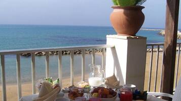 A table with breakfast items, a pitcher of milk, and a potted plant on a balcony overlooking the sea.