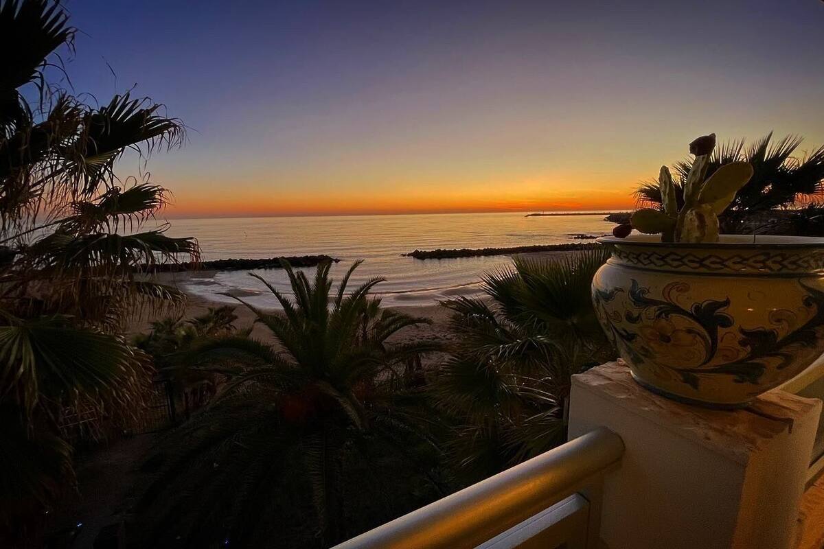 A balcony with a view of the sunset over the ocean, palm trees, and a decorative ceramic pot.