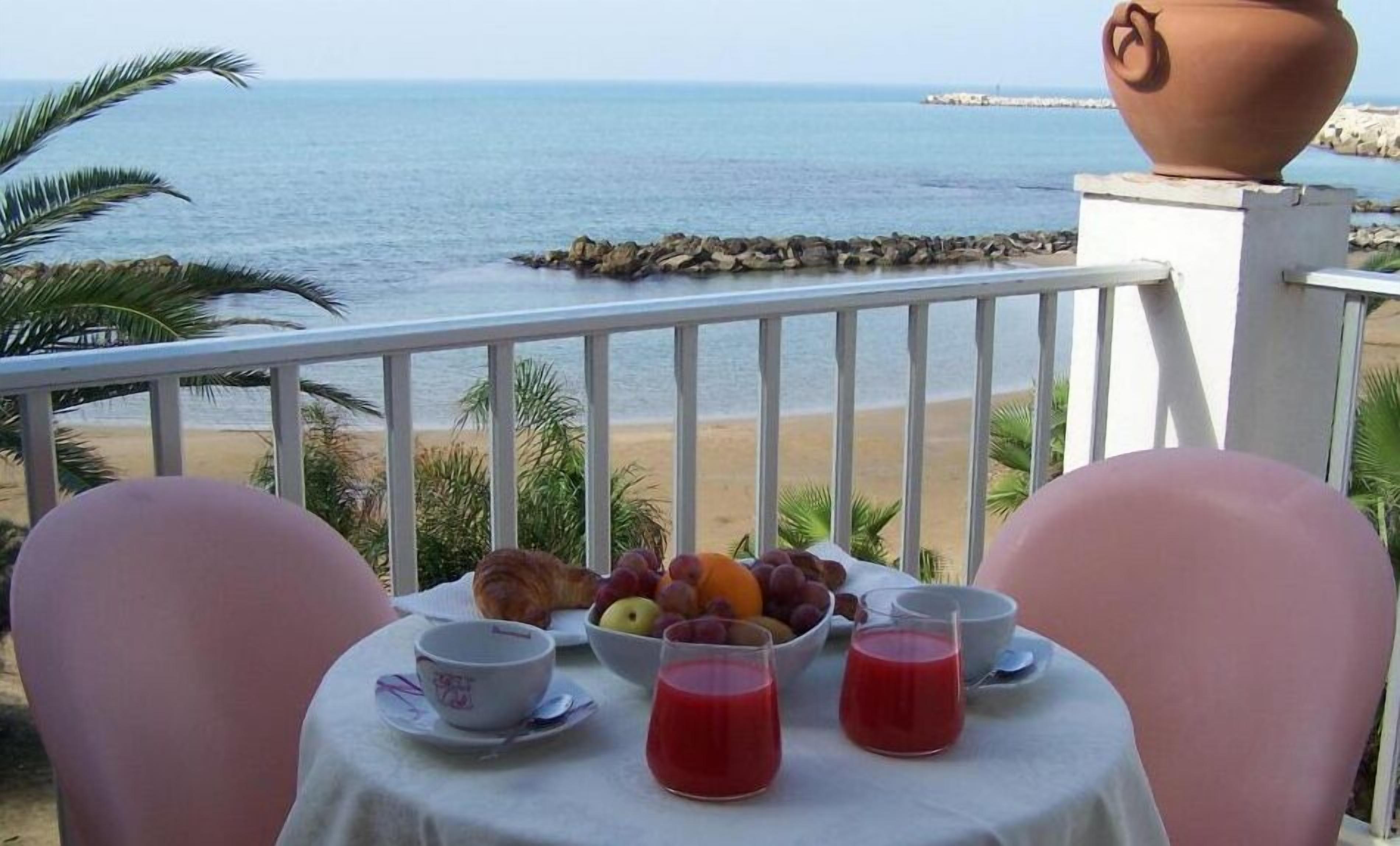 A table with a fruit bowl, two cups, and two glasses of red drink, overlooking a beach and the sea.