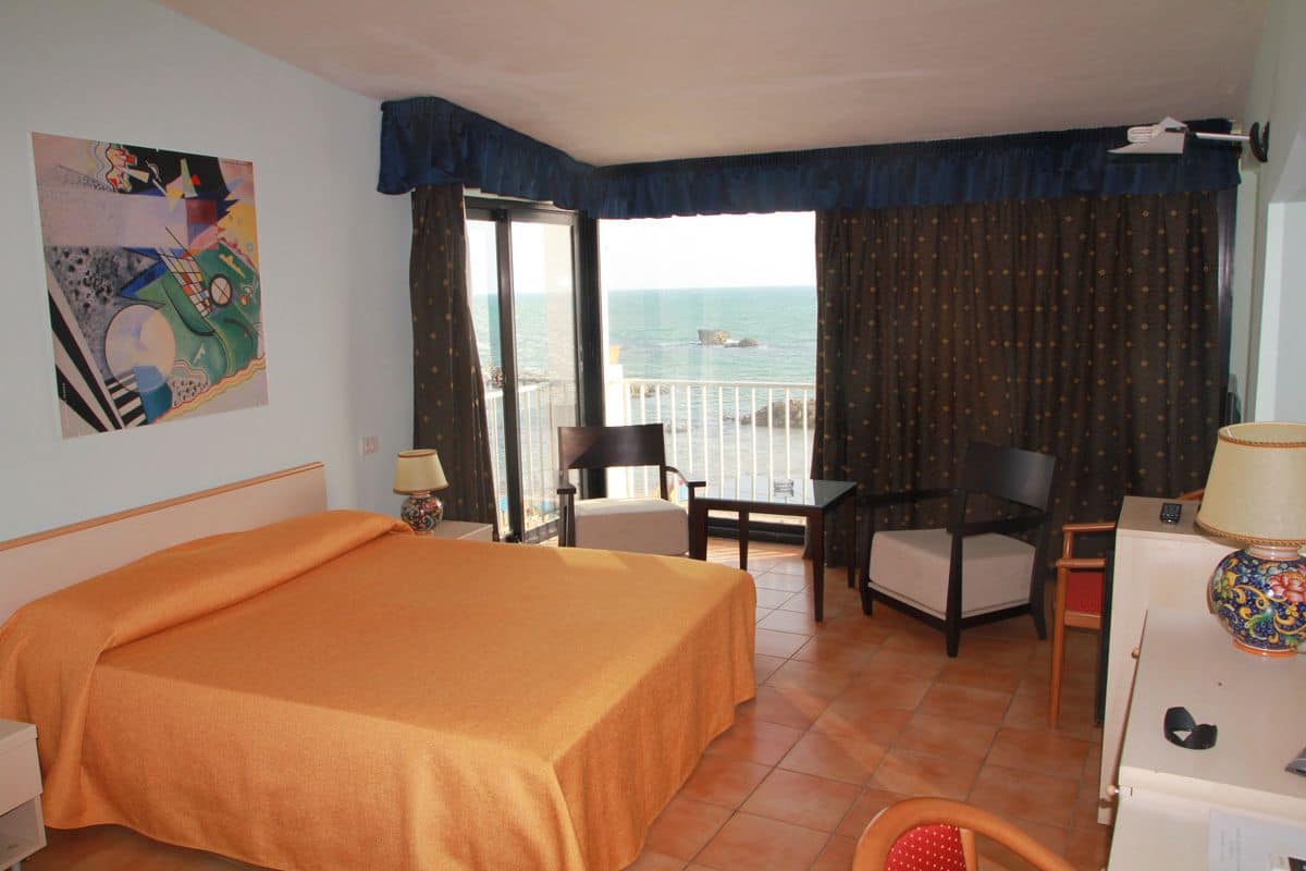 A hotel room with a bed, a desk, two chairs, a lamp, a painting, and a view of the sea.