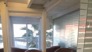 Un espacio interior moderno con un ventanal que da a un balcón con vistas a palmeras y una piscina. Hay varias sillas marrones y una pequeña mesa redonda.