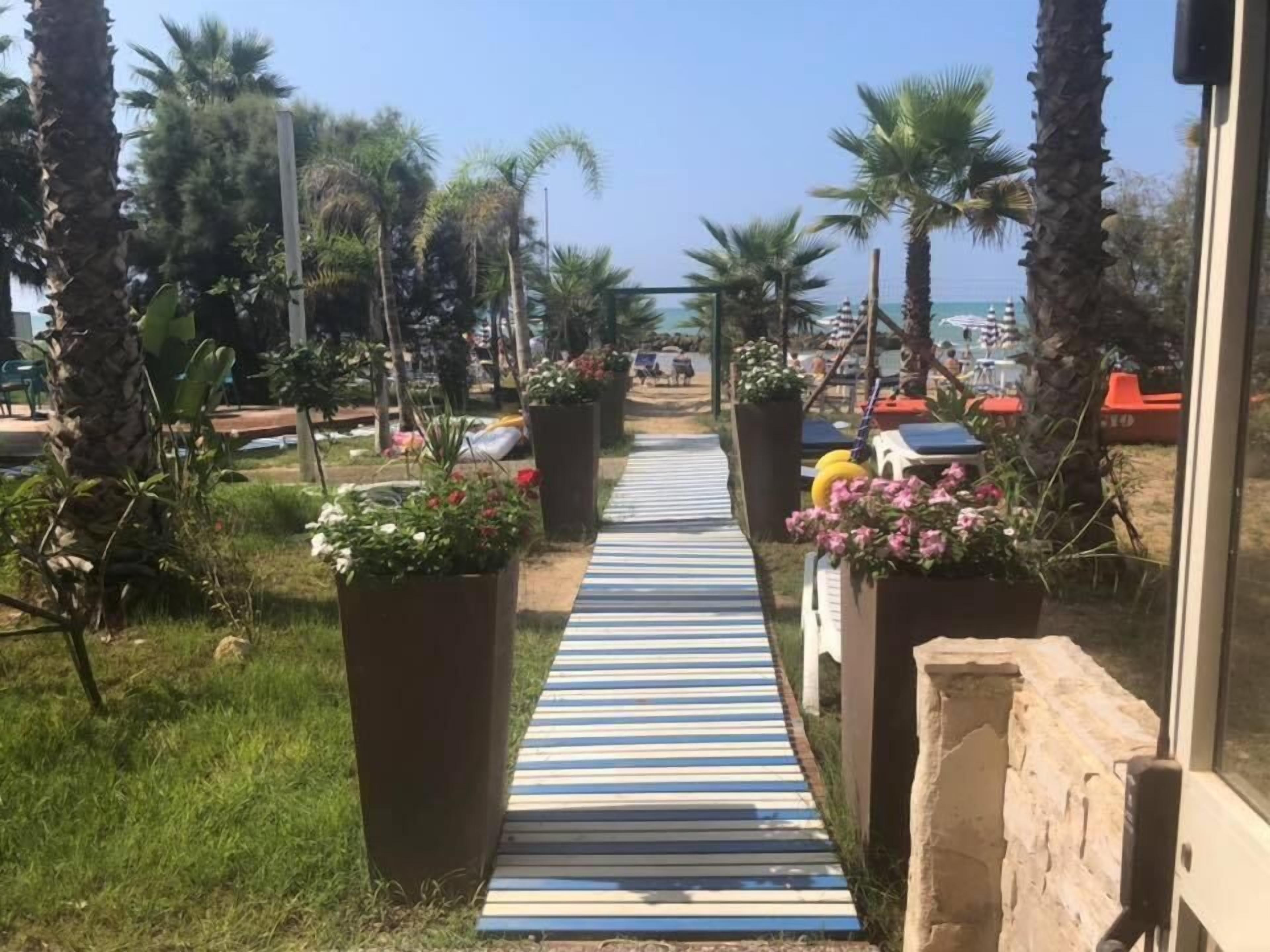 Ein Weg führt zu einem Strandbereich mit Palmen und Topfpflanzen.