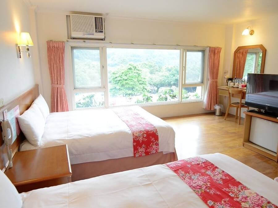 A hotel room with two beds, a desk with a television, and large windows with curtains.