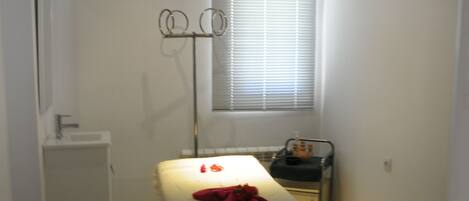 A small, narrow room with a single bed, a window with blinds, a radiator, and a small table with a chair.
