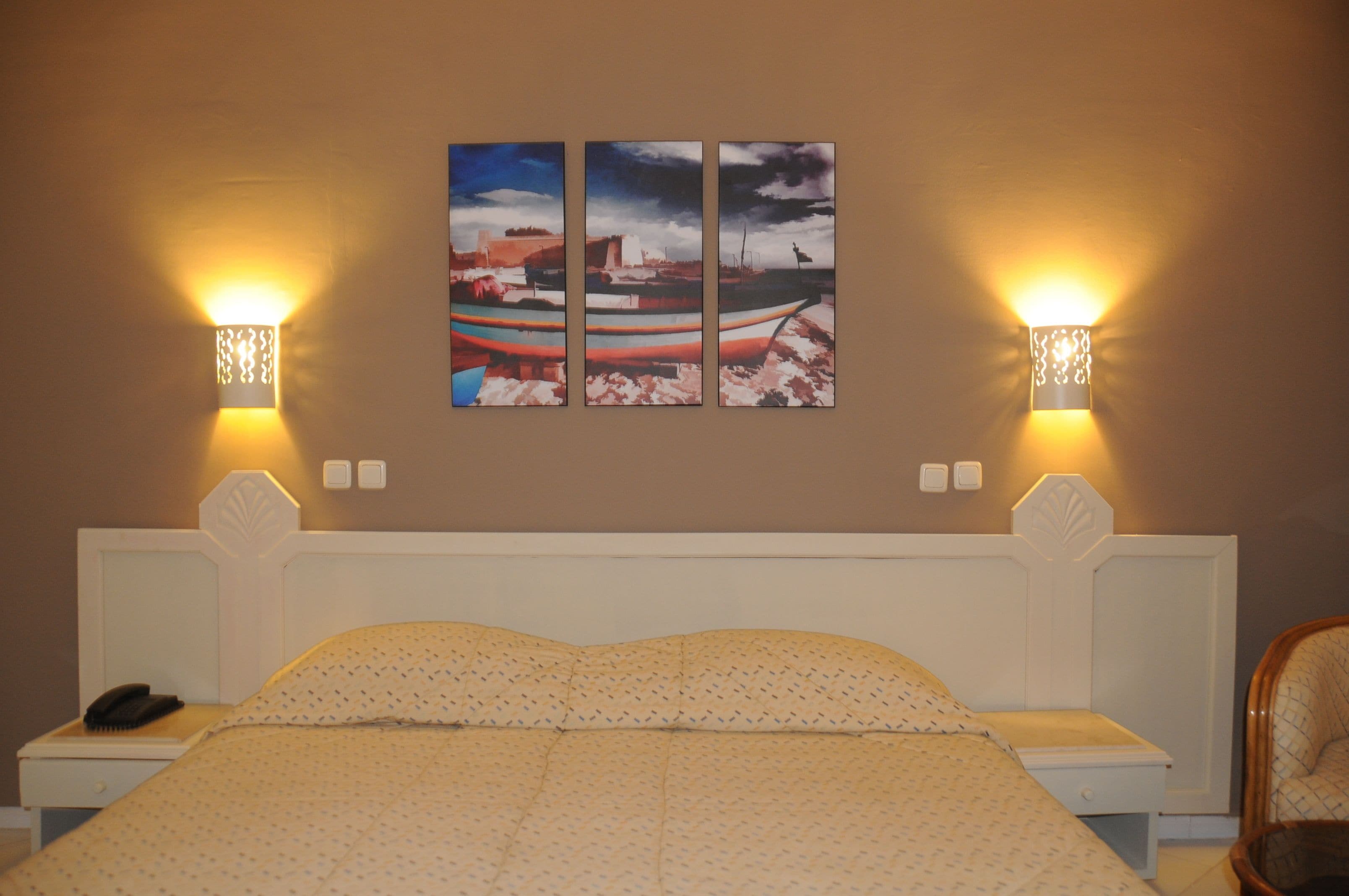 Habitación de hotel con cama, dos mesitas de noche, una silla y un cuadro en la pared que representa un barco.