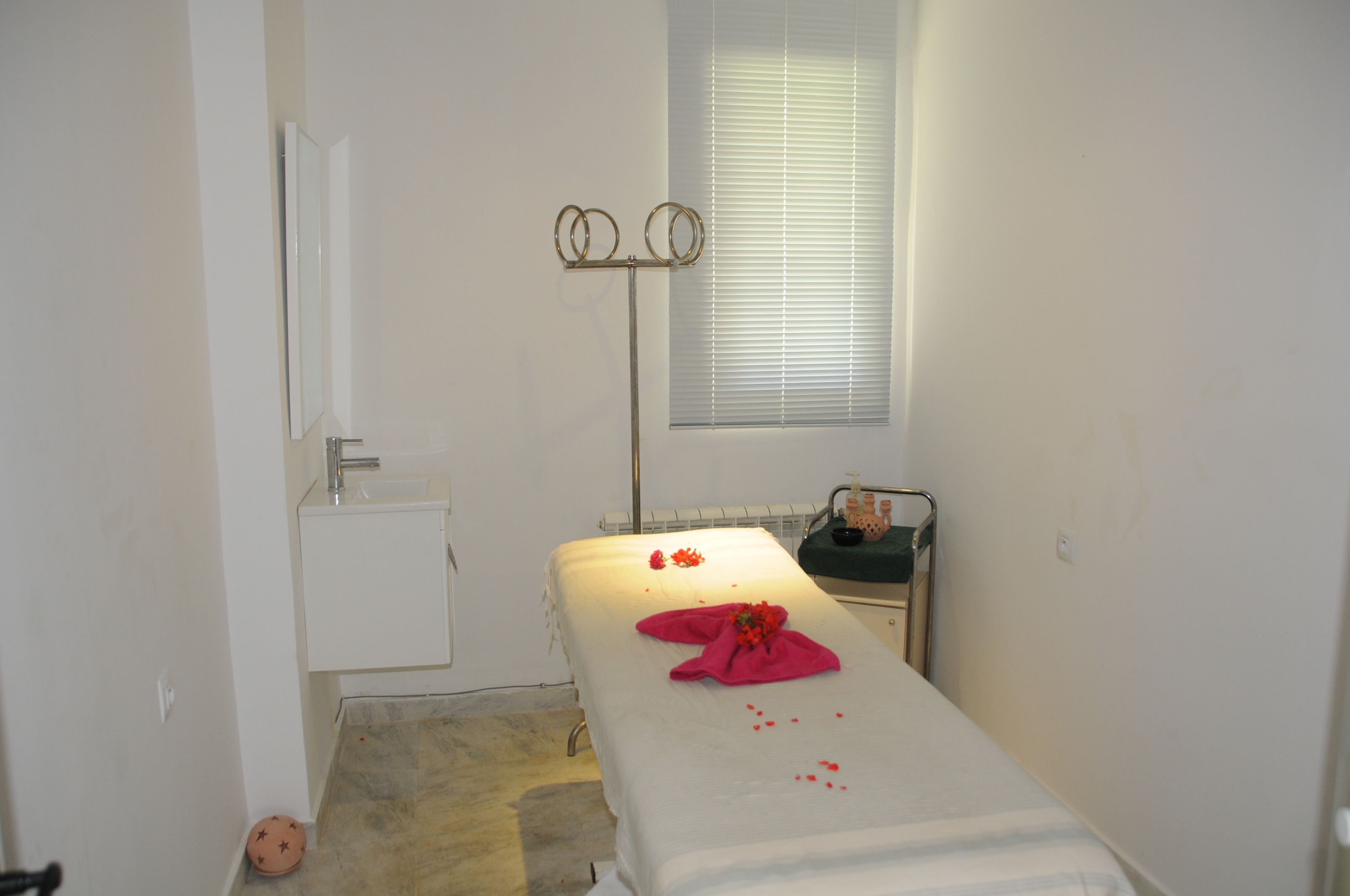 A small, clean room with a single bed, a small table with toiletries, a mirror, and a window with blinds.