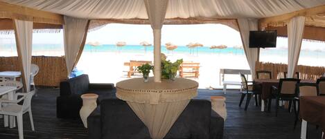 An indoor dining area with a view of the beach, featuring tables and chairs, a central table with a plant, and a TV screen.