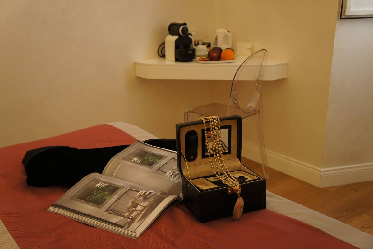 A room with a bed, a bedside table with a book, a clear jewelry box with a necklace, and a shelf with a coffee maker and fruit.