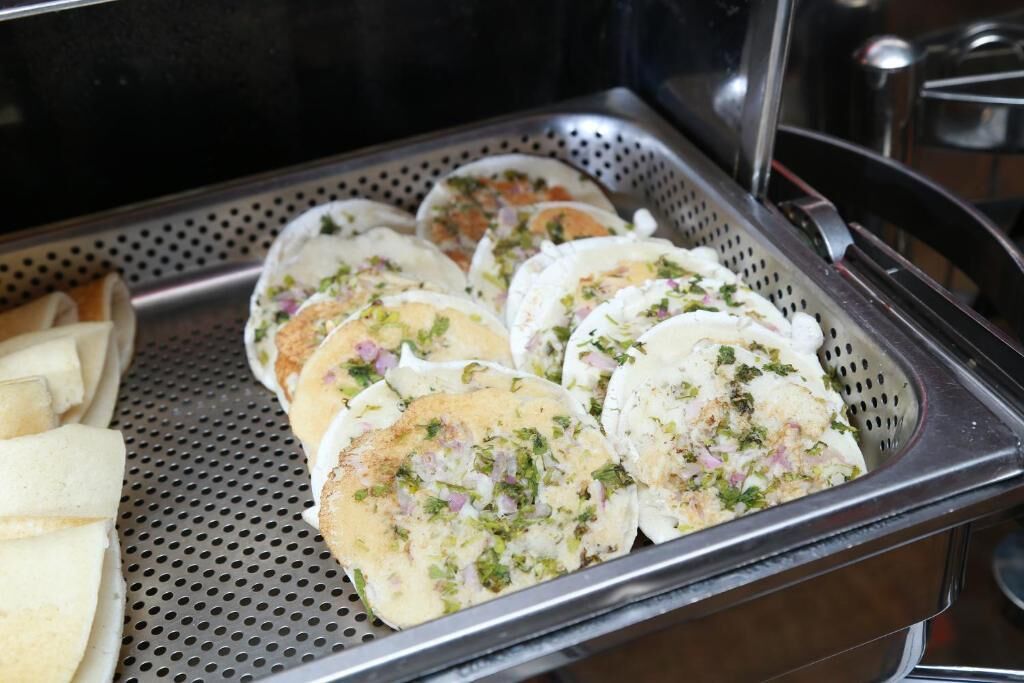 A tray of food items, likely a type of flatbread or pancake, garnished with herbs and onions, placed on a perforated metal tray.