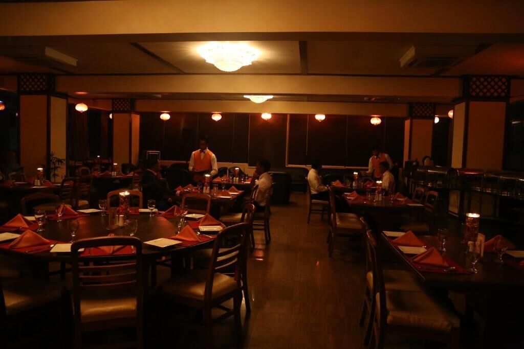 A dimly lit restaurant with round tables and chairs, a chandelier, and a bar area.