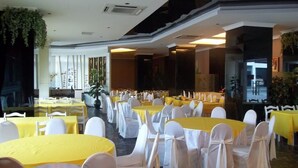 Uma sala de banquetes com mesas redondas, cadeiras brancas e toalhas amarelas.