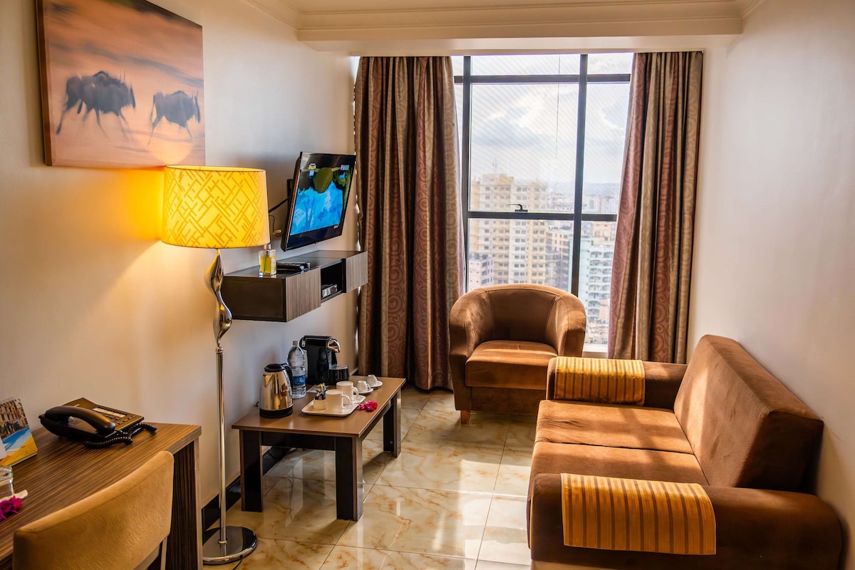 A hotel room with a sofa, a coffee table, a TV, a lamp, and a window with curtains.