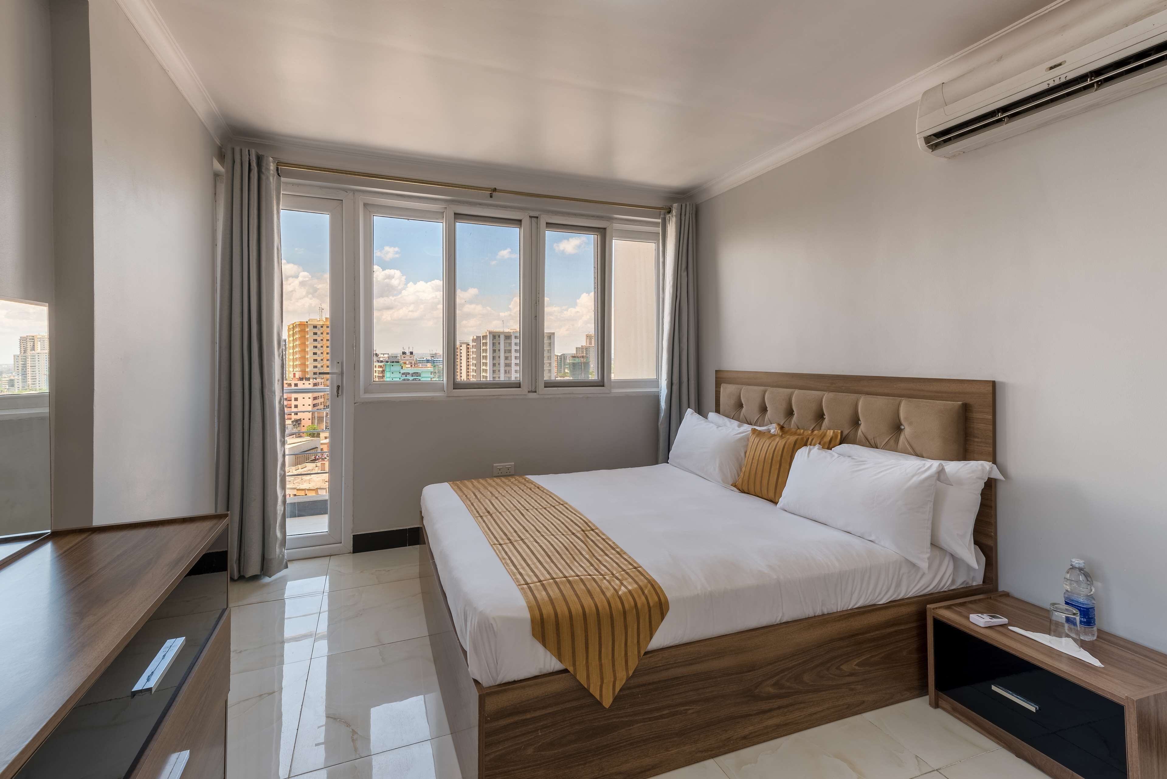 A hotel room with a large bed, a wooden headboard, a bedside table, and a view of the city through large windows.