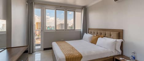 A hotel room with a large bed, a wooden headboard, a bedside table, and a view of the city through large windows.