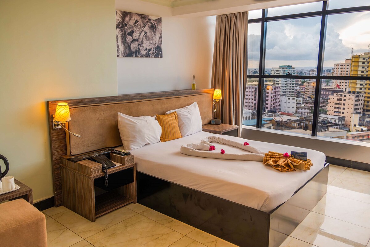 A hotel room with a large bed, a city view, a painting on the wall, and a lamp on the nightstand.