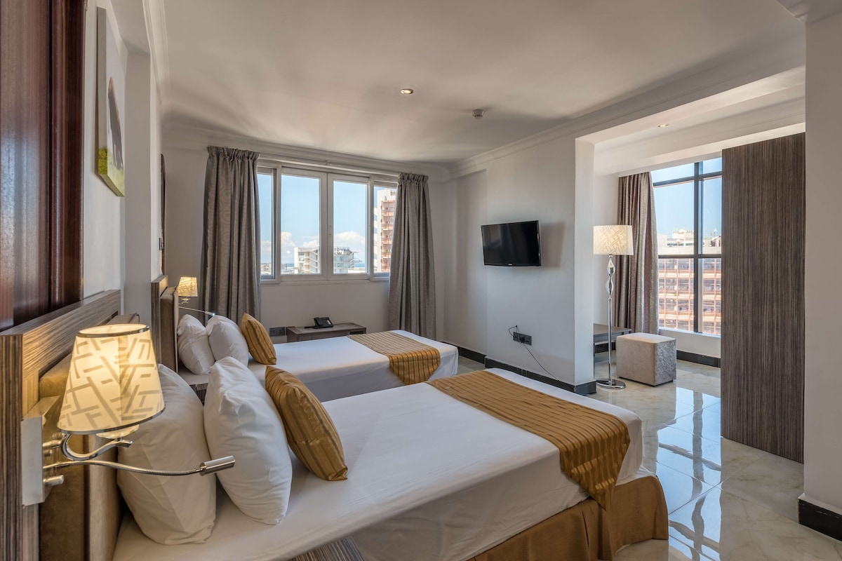 A hotel room with two beds, a flat-screen TV, a lamp, and a view of the city.