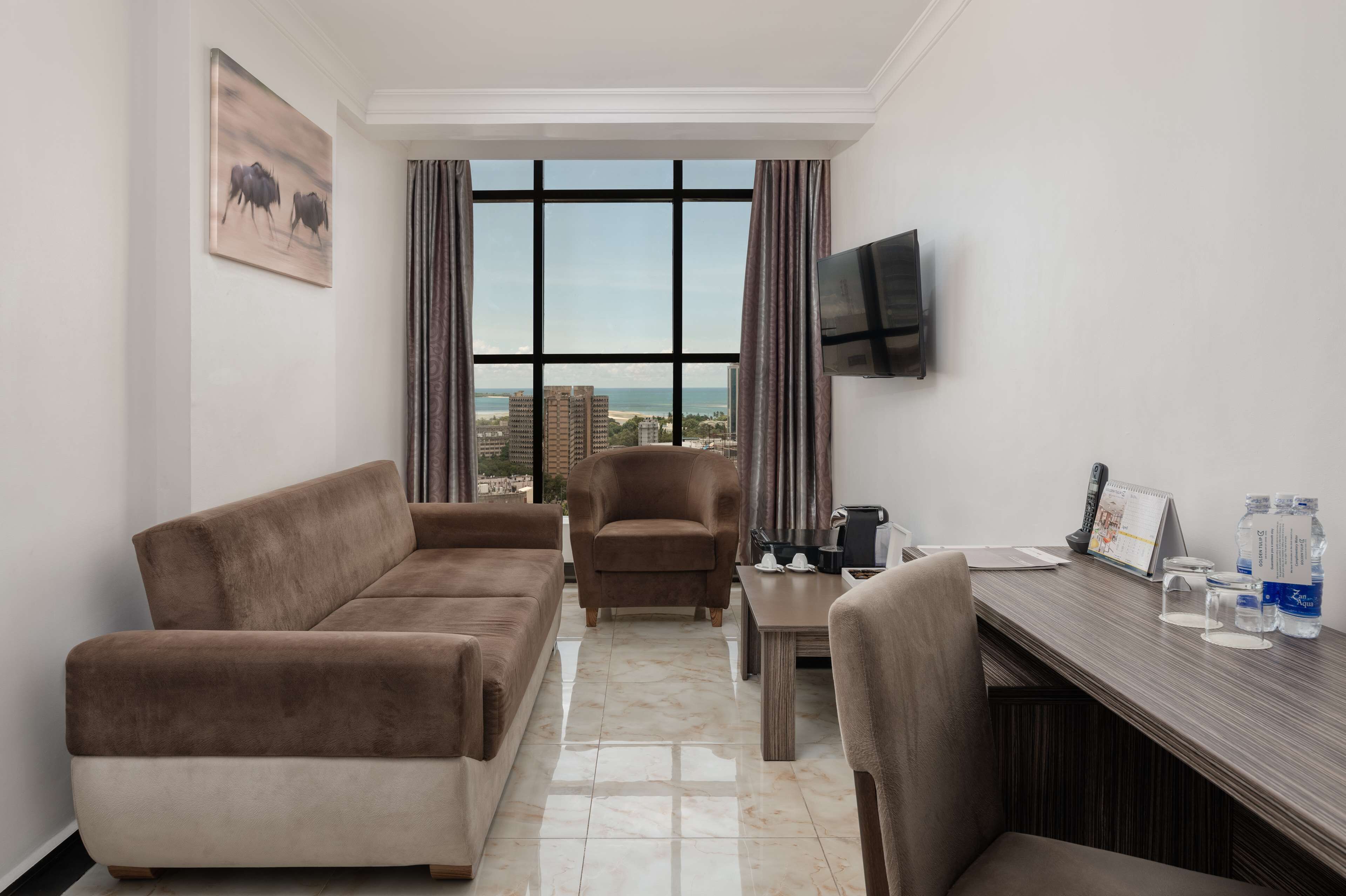 Une chambre d’hôtel dotée d’une grande fenêtre, d’un canapé, d’un fauteuil, d’un bureau et d’un téléviseur à écran plat.