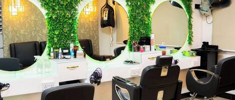 Un salón de belleza con tres sillas negras, una barra blanca con espejos y una pared cubierta de follaje verde.