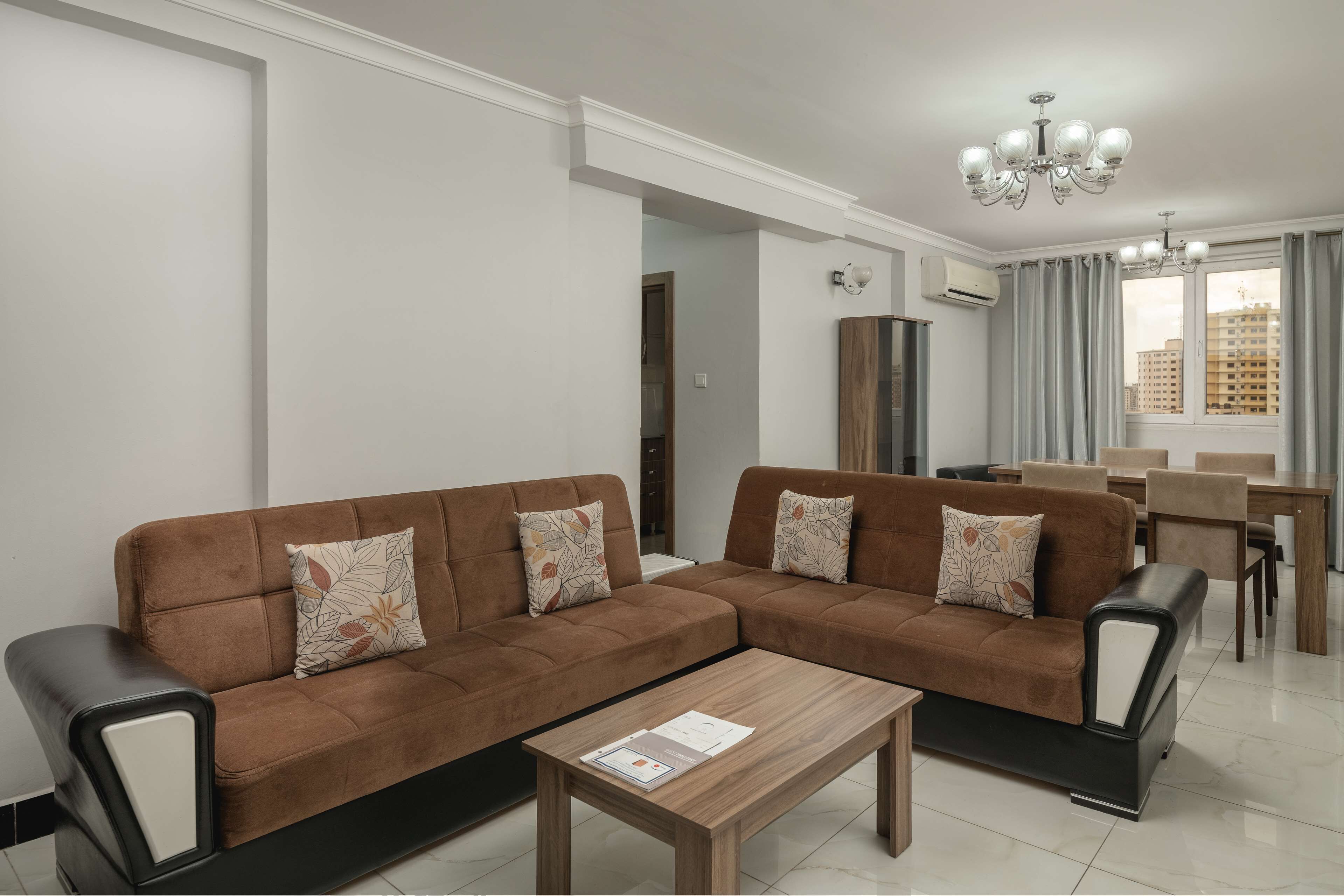 A modern living room with a brown sectional sofa, a wooden coffee table, and a dining area with a chandelier.