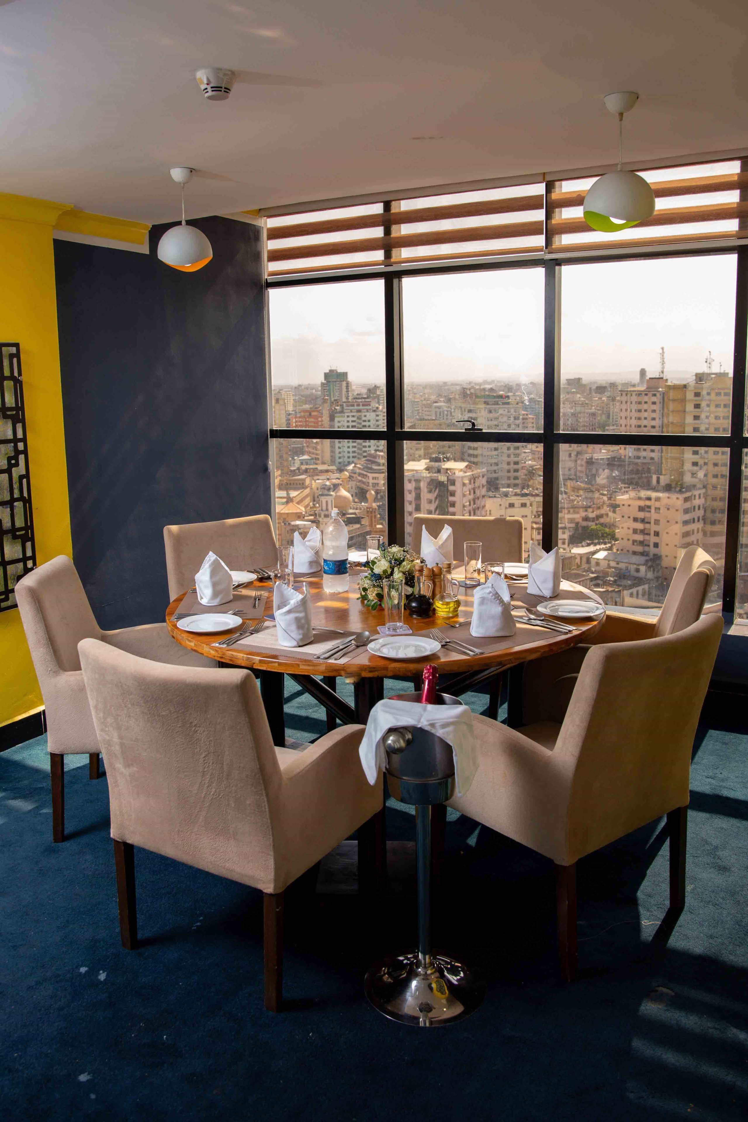 A dining area with a round table set for four, large windows with a city view, and modern pendant lighting.