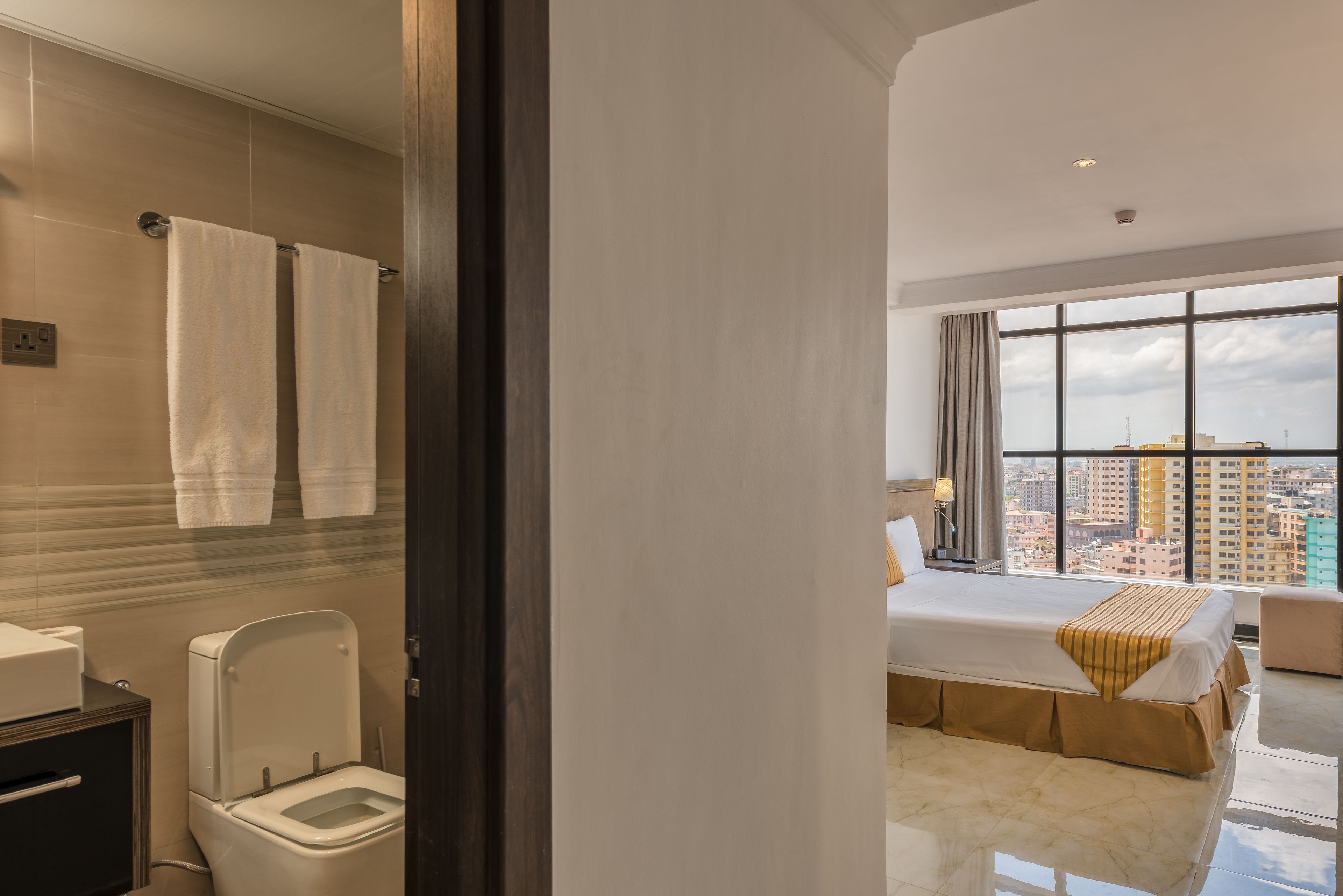 Quarto de hotel moderno com uma cama grande, vista da cidade através de uma janela ampla e banheiro com vaso sanitário e porta-toalhas.