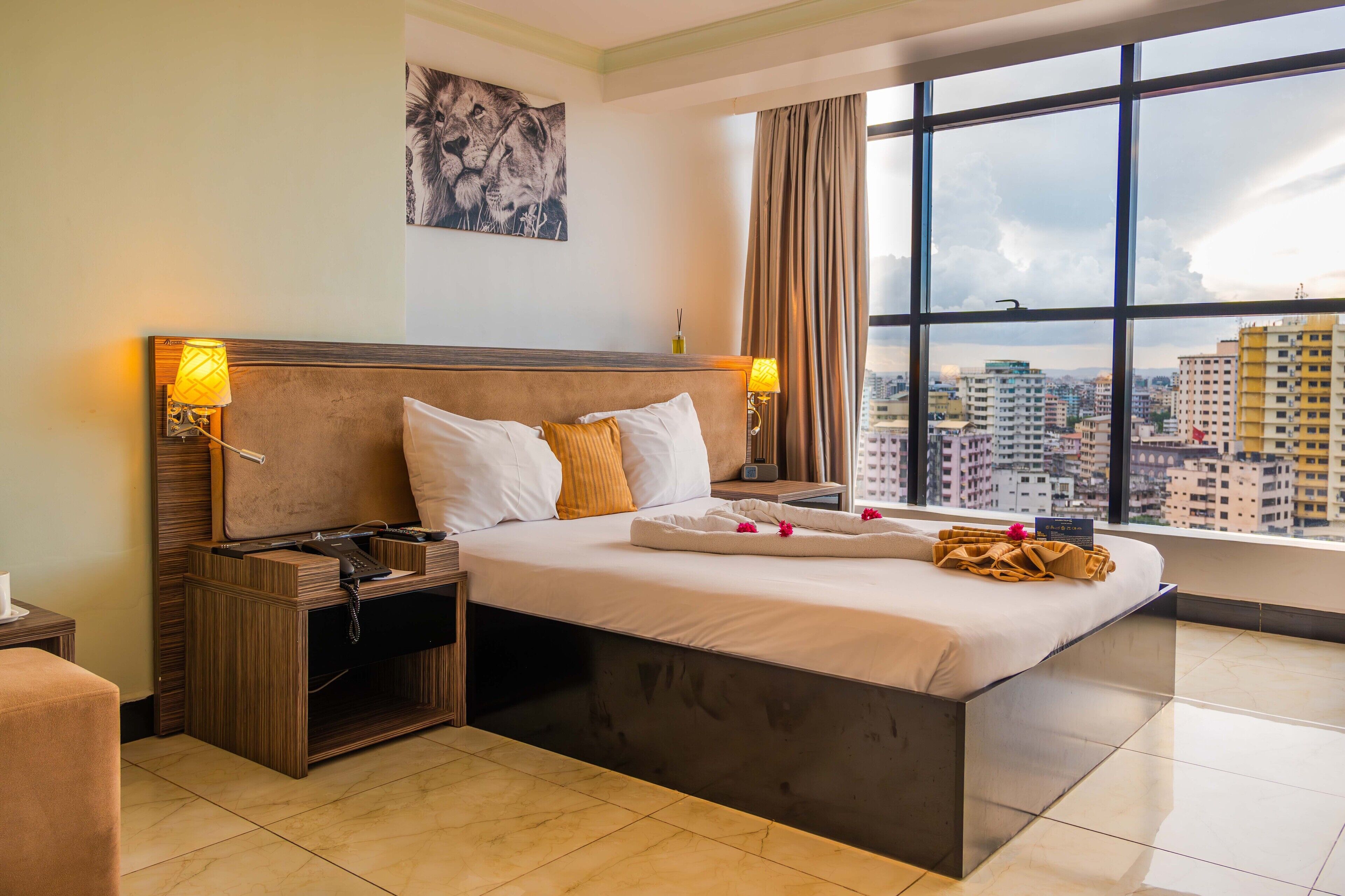 Habitación de hotel con una cama grande, lámparas de noche, una mesita de noche, vistas de la ciudad a través de amplias ventanas y un cuadro en la pared.