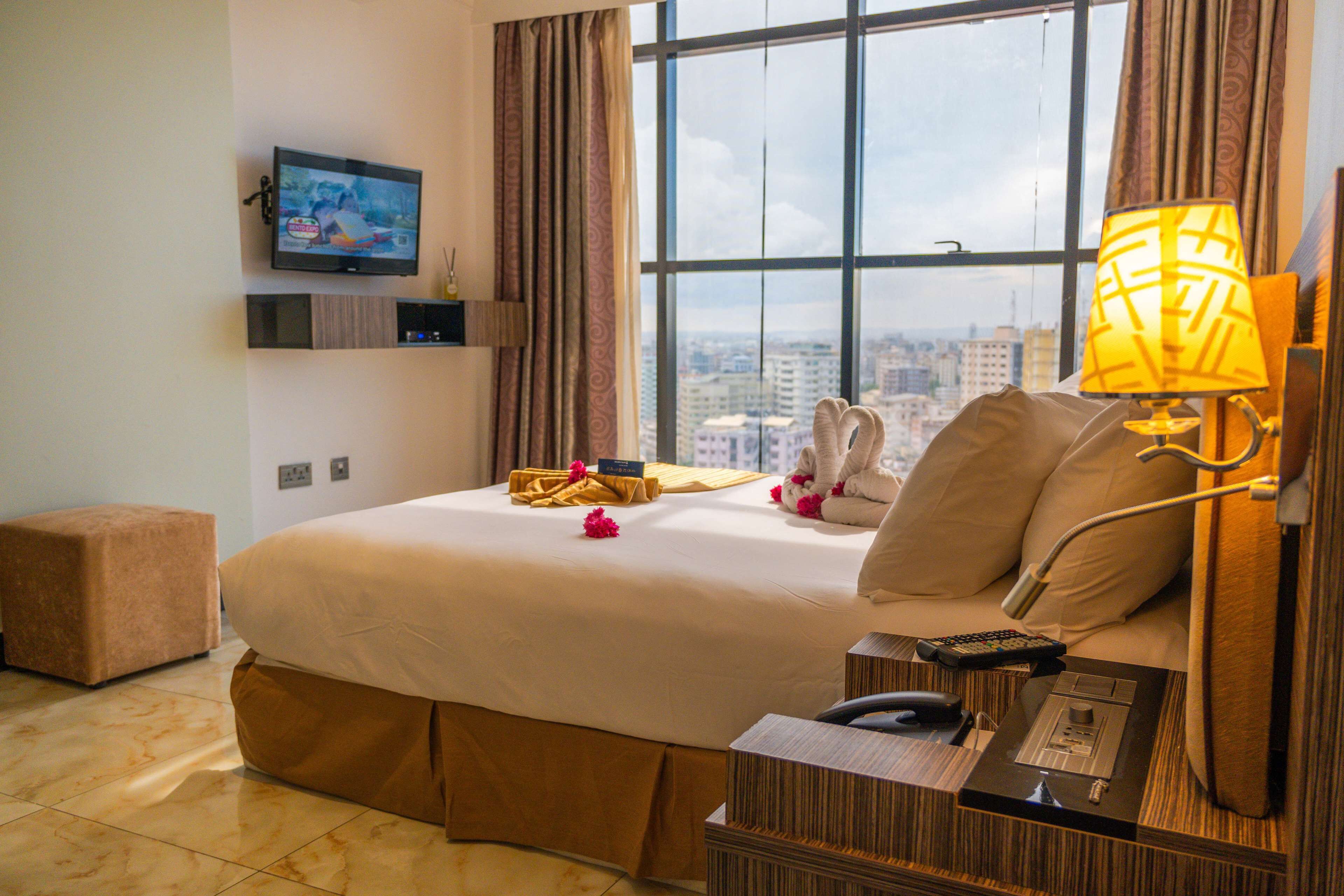 Habitación de hotel con una cama grande, un televisor en la pared, una mesita de noche con lámpara y vistas a la ciudad por la ventana.