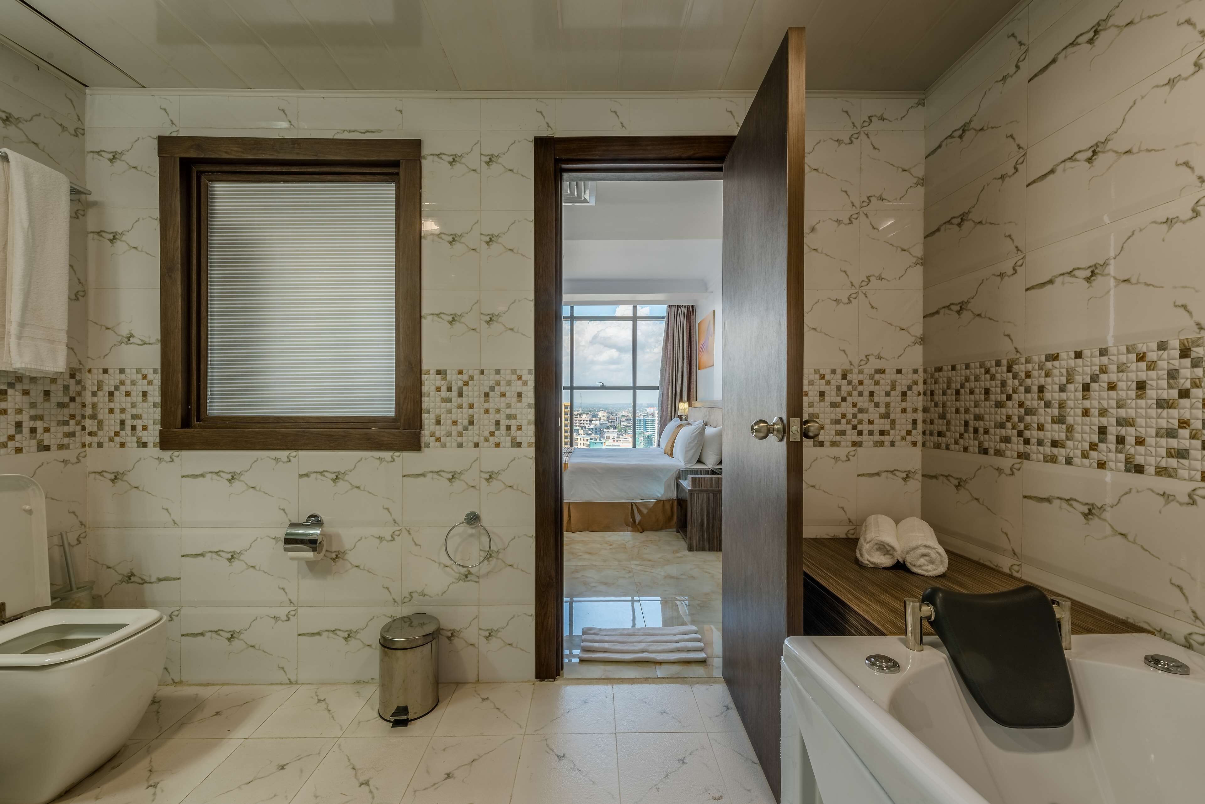 Een badkamer met toilet, bidet en badkuip. Er is een raam met zonwering, een handdoekhouder en een prullenbak.