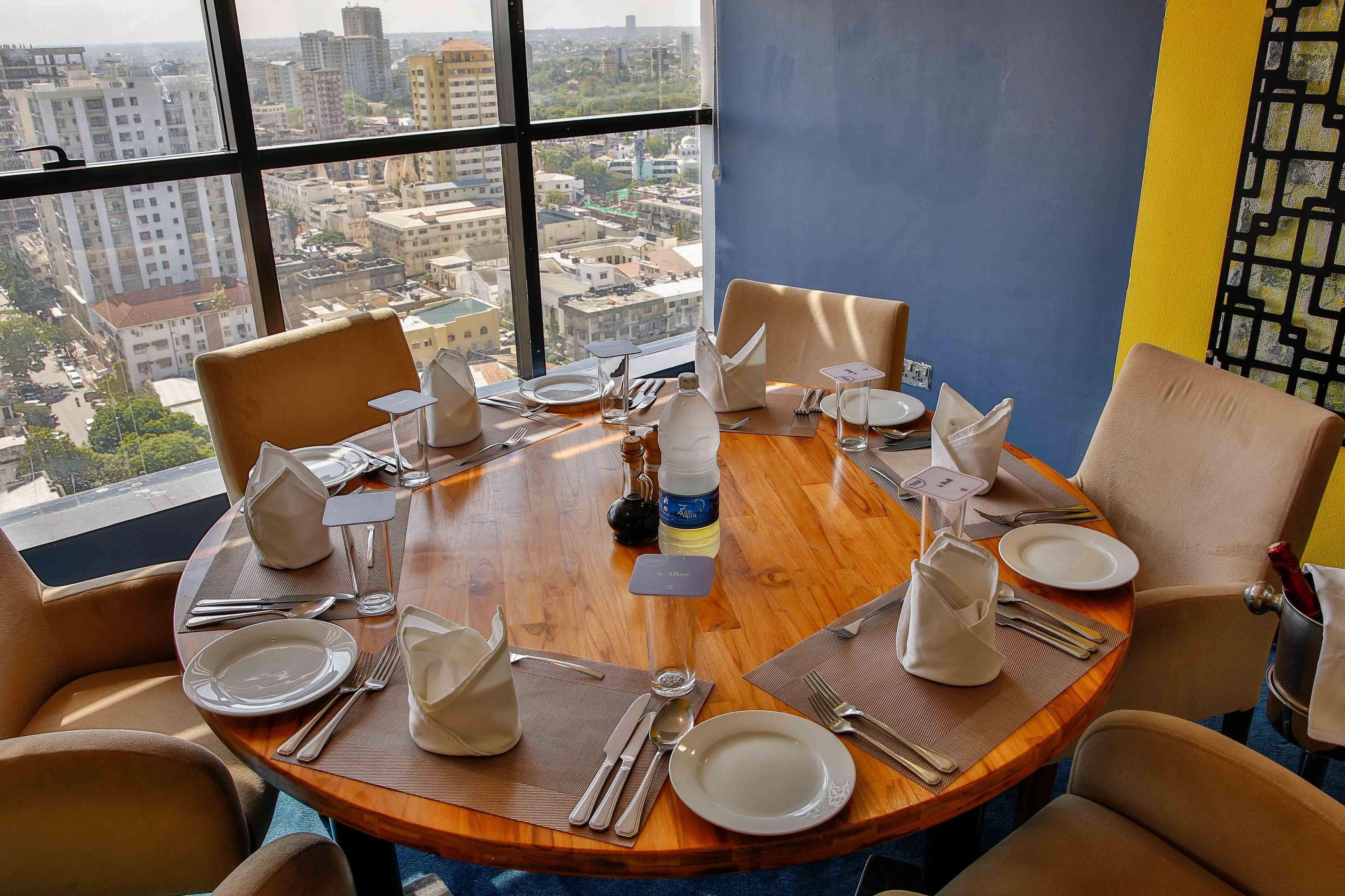 Une table de salle à manger ronde, entourée de chaises, est dressée avec de l’argenterie, des couverts et de la vaisselle blanche. De grandes fenêtres offrent une vue sur la ville.