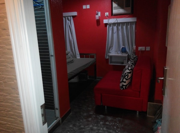 A compact room with a red wall, a small bed, a red sofa, a window with curtains, and a wall-mounted air conditioner. - Blessing Guesthouse (Kowloon)
