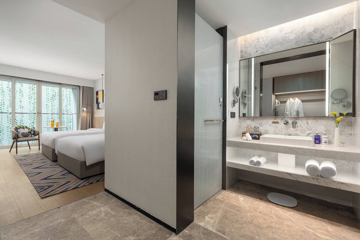 Une chambre d’hôtel moderne dotée d’un grand lit, d’une salle de bain séparée avec un meuble-lavabo et offrant une vue sur la verdure par la fenêtre.