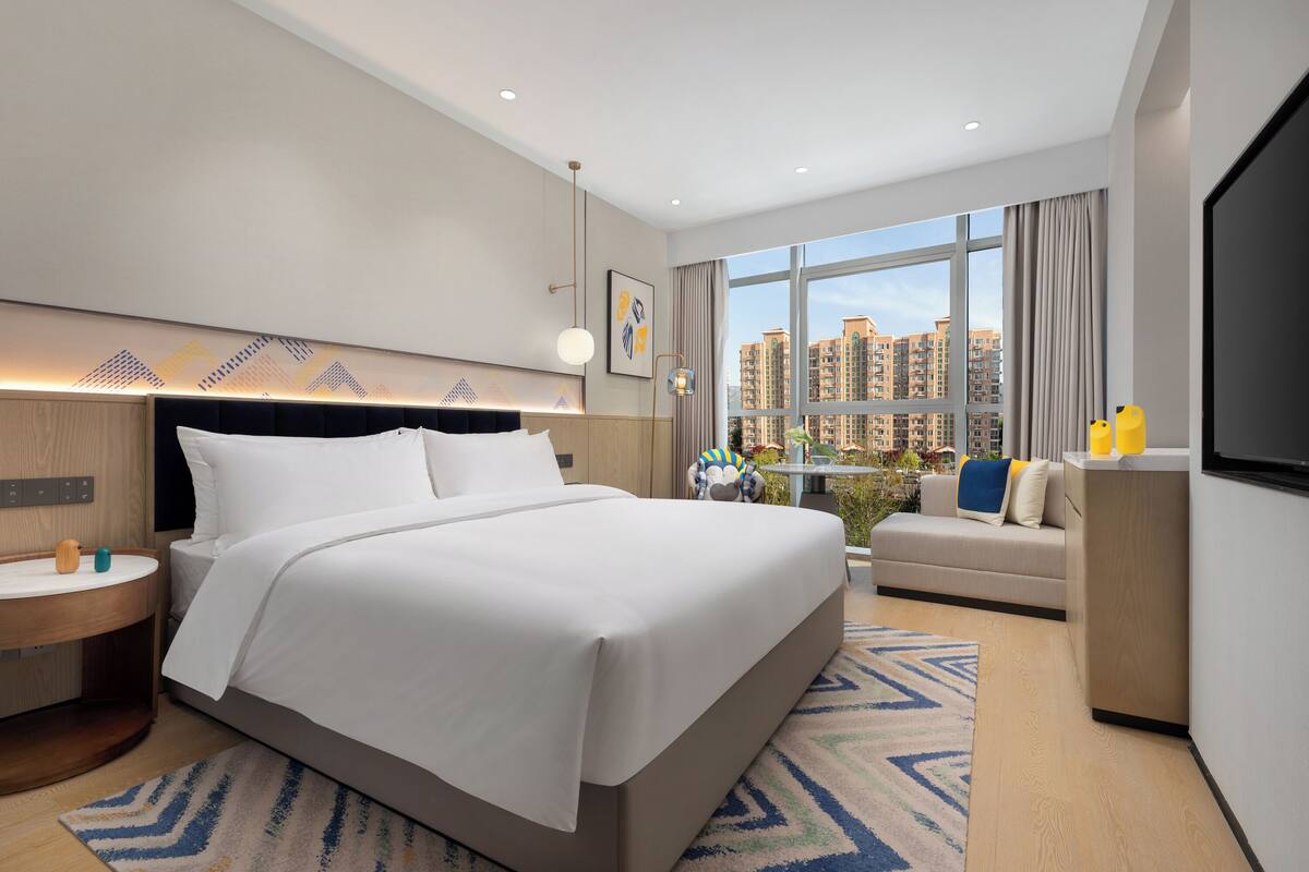 Ein modernes Hotelzimmer mit einem großen Bett, einer Couch, einem Fernseher und Blick auf hohe Gebäude.