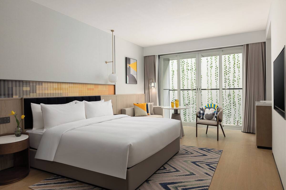 Ein Hotelzimmer mit einem großen Bett, einem Nachttisch mit Blumenvase, einem Sessel, einem kleinen Tisch mit Getränken und einem Fernseher.
