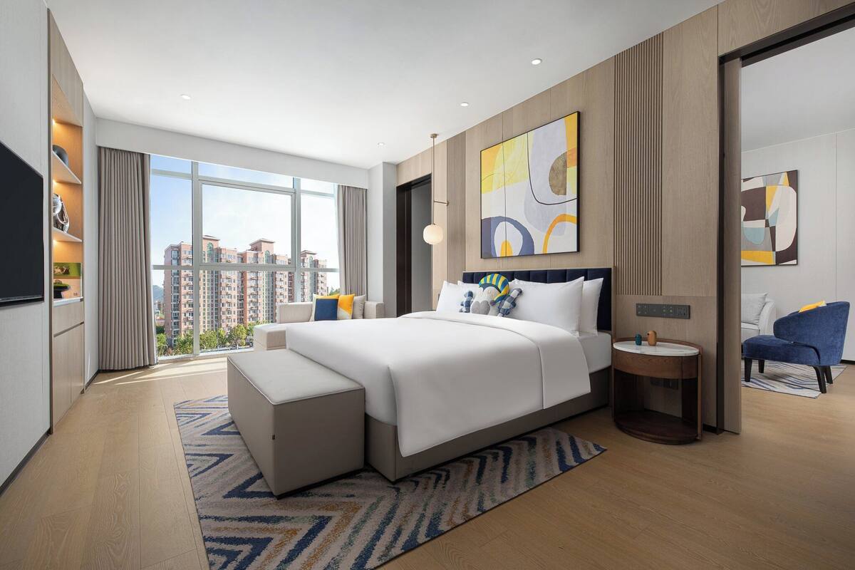 Ein modernes Hotelzimmer mit einem großen Bett, einem Nachttisch, einem Fernseher und Blick auf die Stadt.