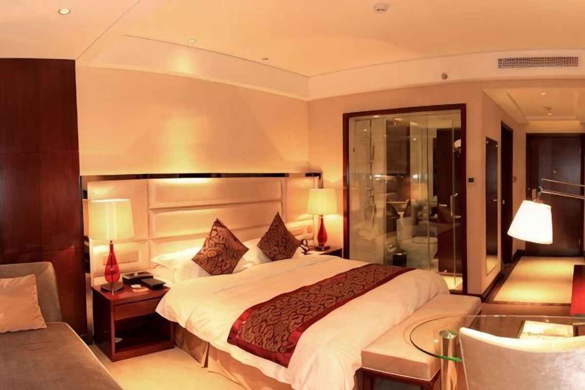 Ein Hotelzimmer mit einem großen Bett, Nachttischlampen, einer Glastür zum Badezimmer und einem kleinen Tisch mit Stuhl.