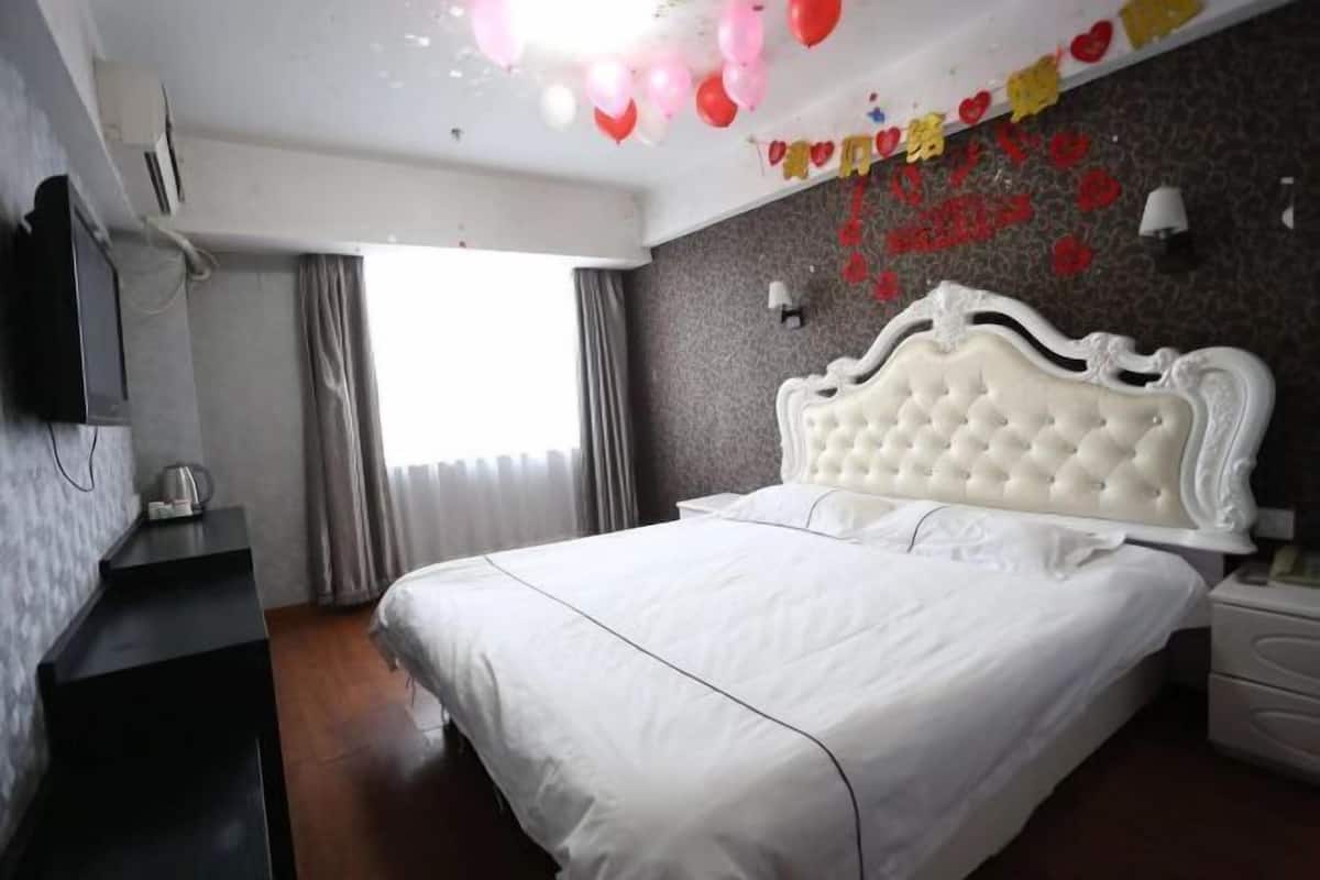 A hotel room with a white bed, a flat-screen TV, a bedside table, and a window with curtains.
