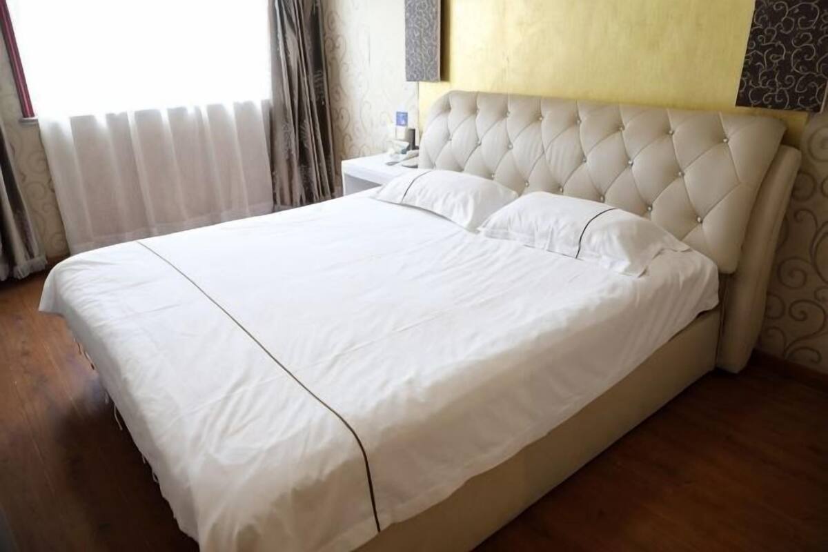 A large bed with a tufted headboard, a bedside table, and a window with curtains.