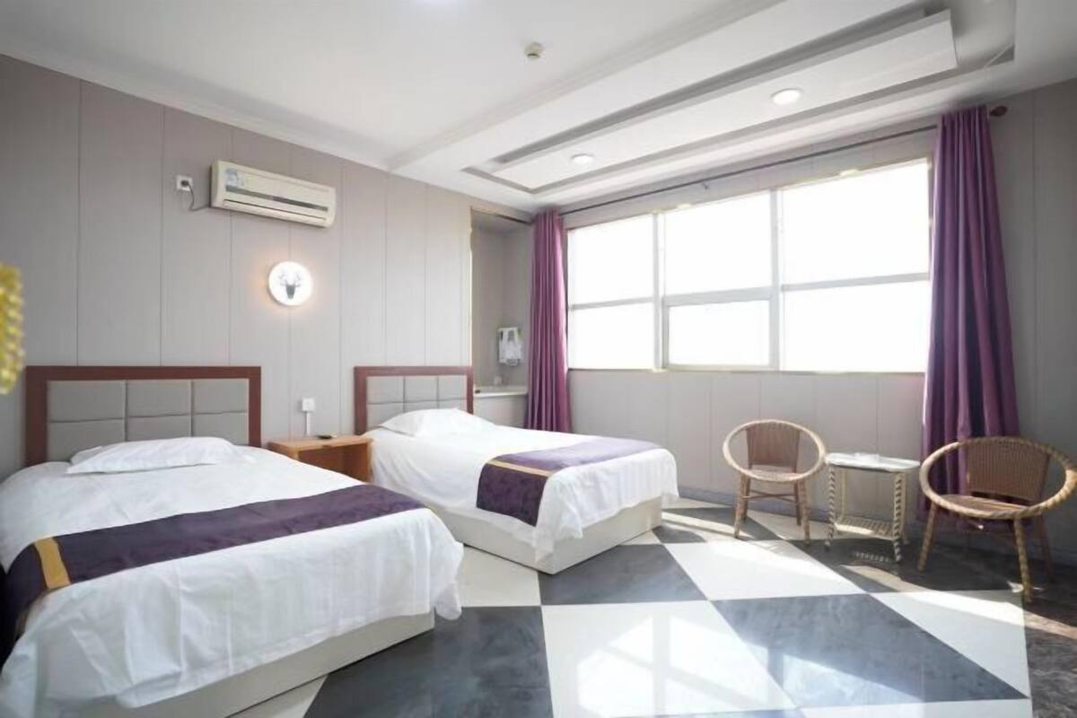 A hotel room with two beds, a window with purple curtains, a small table, and a chair.