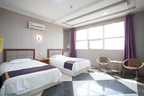 Quarto de hotel com duas camas, uma janela com cortinas roxas, uma mesa pequena e uma cadeira.