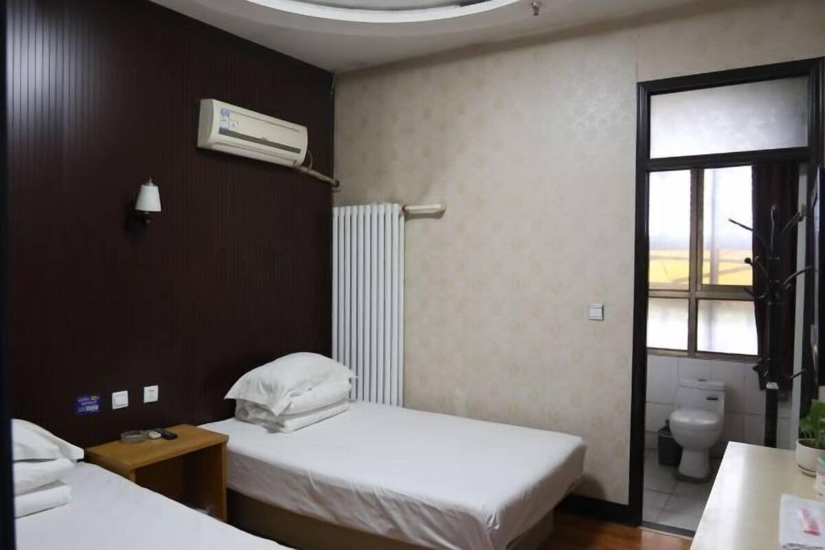 A hotel room with two beds, a window, a radiator, an air conditioner, a bedside table, and a toilet.