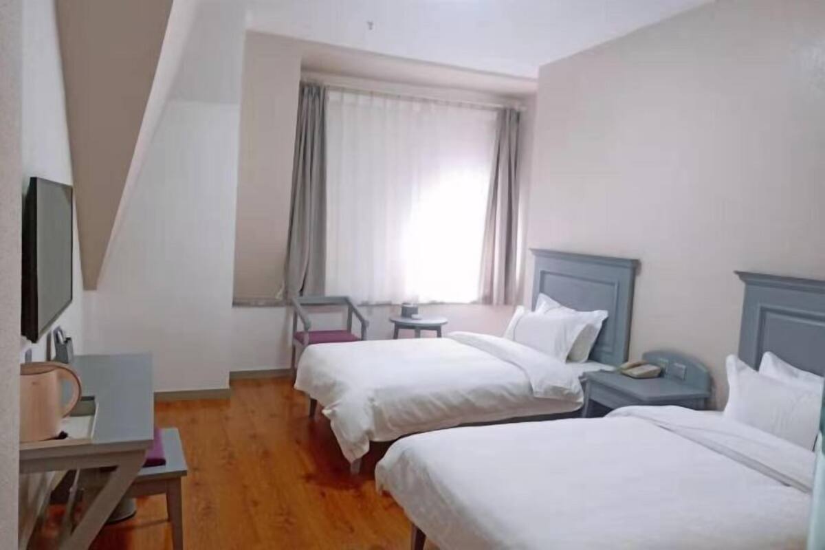 A hotel room with two beds, a small table, a chair, a TV, and a window with curtains.